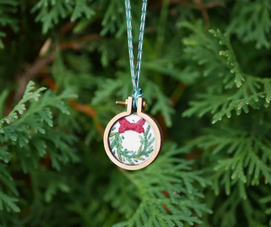 Image of an embroidered wreath Christmas ornament in front of greenery. 