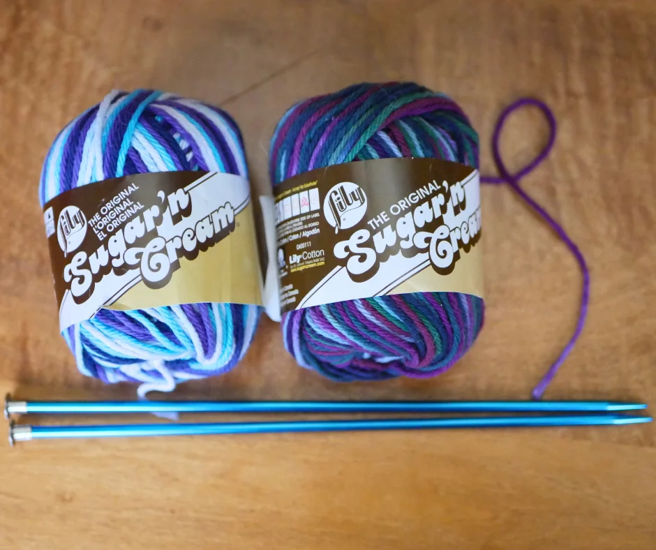 Image of two skeins of cotton yarn in Sugar n Cream brand. The left skein is in blues, whites, and purples. The right skein is in blues, greens, and purples. A pair of blue metal straight knitting needles lay next to the yarn. 