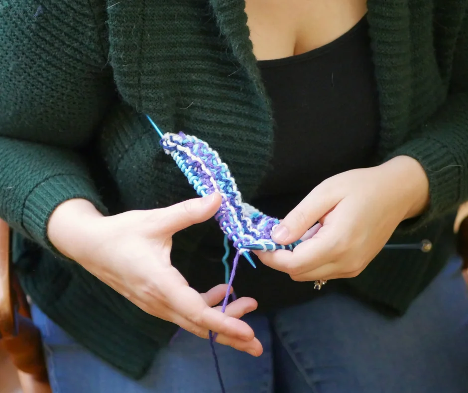 Image of a white person wearing blue jeans, a black shirt, and a green cardigan knitting lever style. 