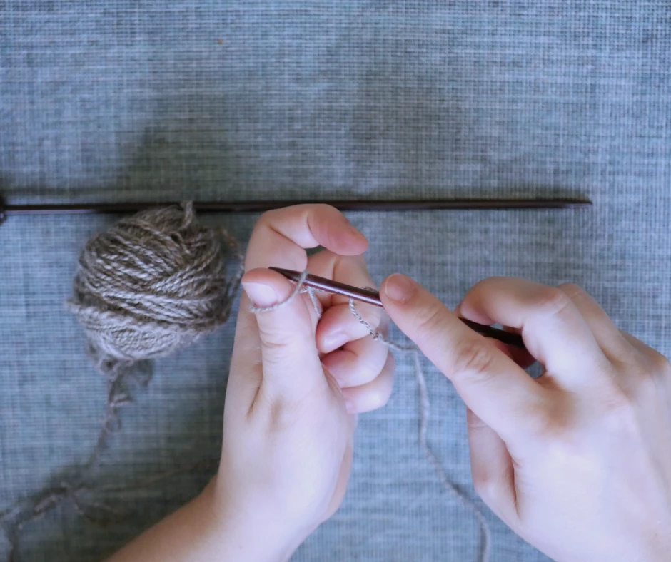Image of a pair of white hands. The right hand is holding a dark wood knitting needle and putting the tip of the knitting needle into a loop of yarn around the left thumb. 