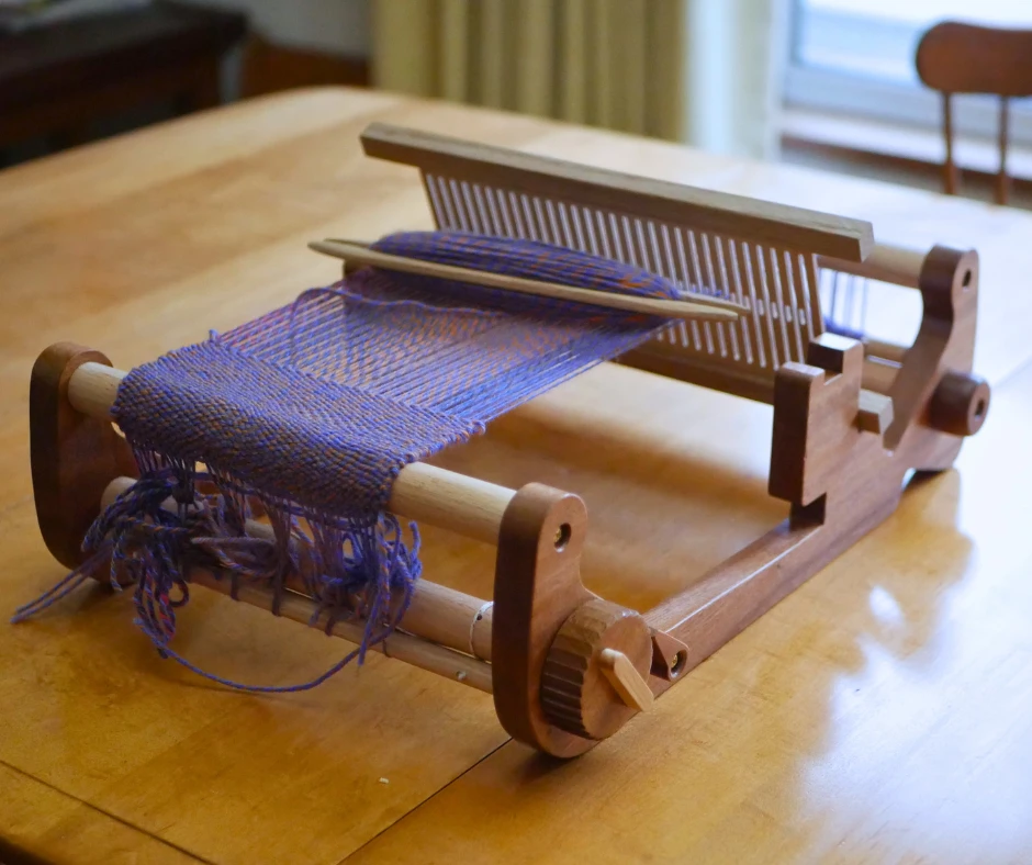 Image of wood rigid heddle table loom with a shuttle full of yarn resting on the warp. 