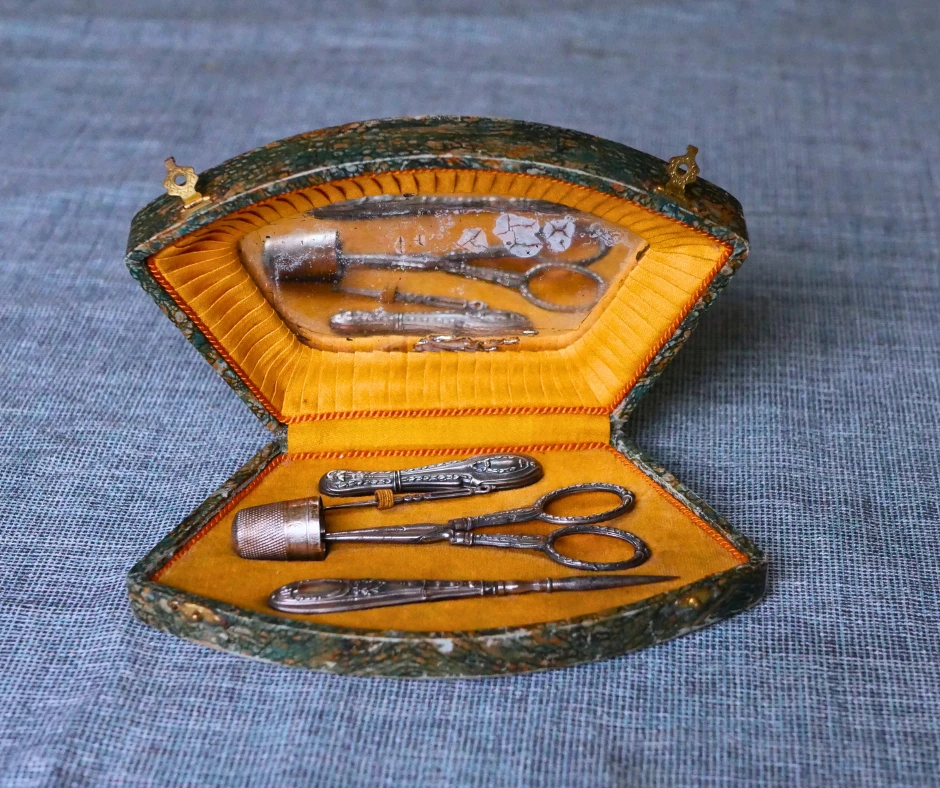 Image of an antique sewing kit that includes a silver awl, thimble, scissors, and needle holder. The outside of the kit is green and the inside is a golden yellow with a mirror in the lid. 