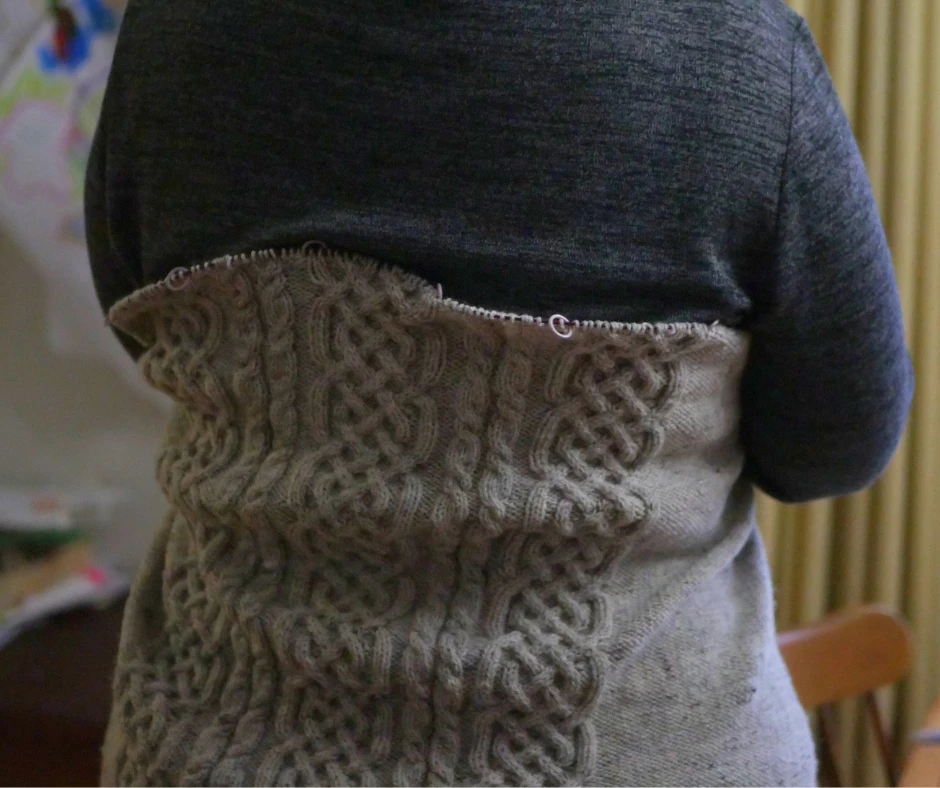 Image of a partially knit cabled sweater wrapped across a woman's back to show how far the back has been knit. 