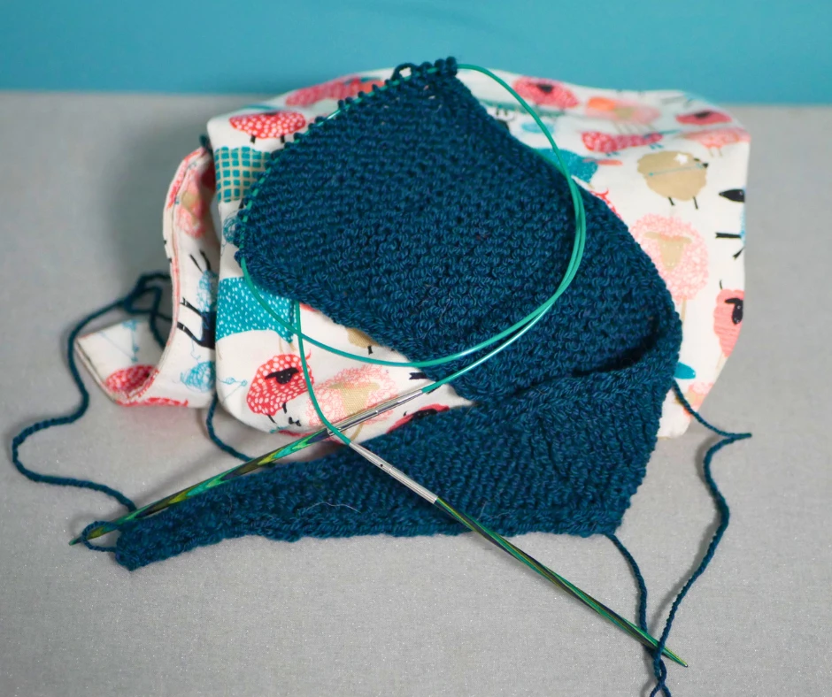 Image of a partially knit Sophie scarf resting on a project bag with a sheep pattern. The yarn, knitting needles, cables and some of the sheep are all teal. 