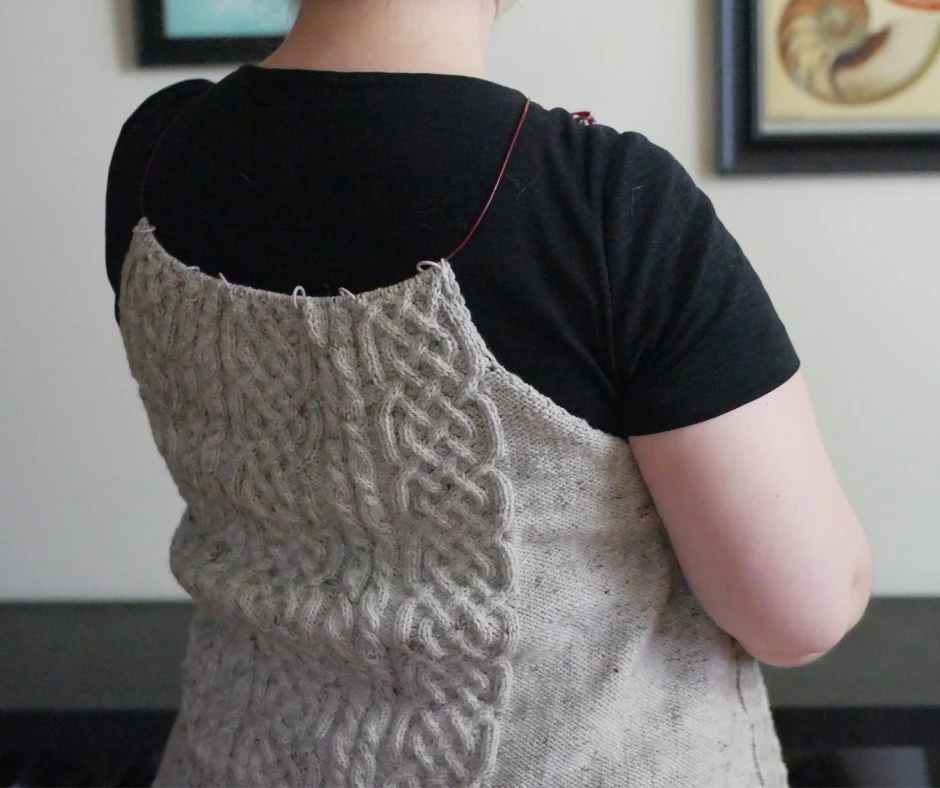 Image of a partially knit cable cardigan being held up to the knitter's back to indicate how far it has been knit.  