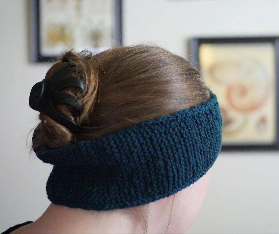 Image of a partially knit scarf wrapped around the knitter's ears so that it does not interfere with her hair pulled up with a claw clip. 