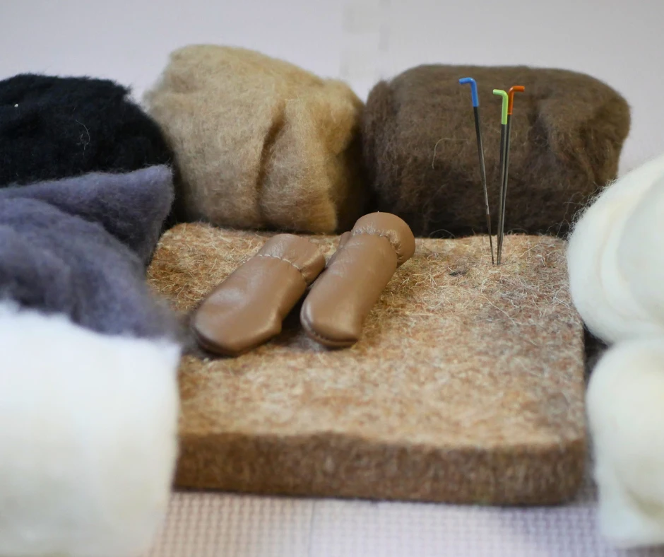 Image of needle felting supplies including a mat to work on, three sizes of felting needles, a set of leather finger guards, and felting wool in a variety of colors. 
