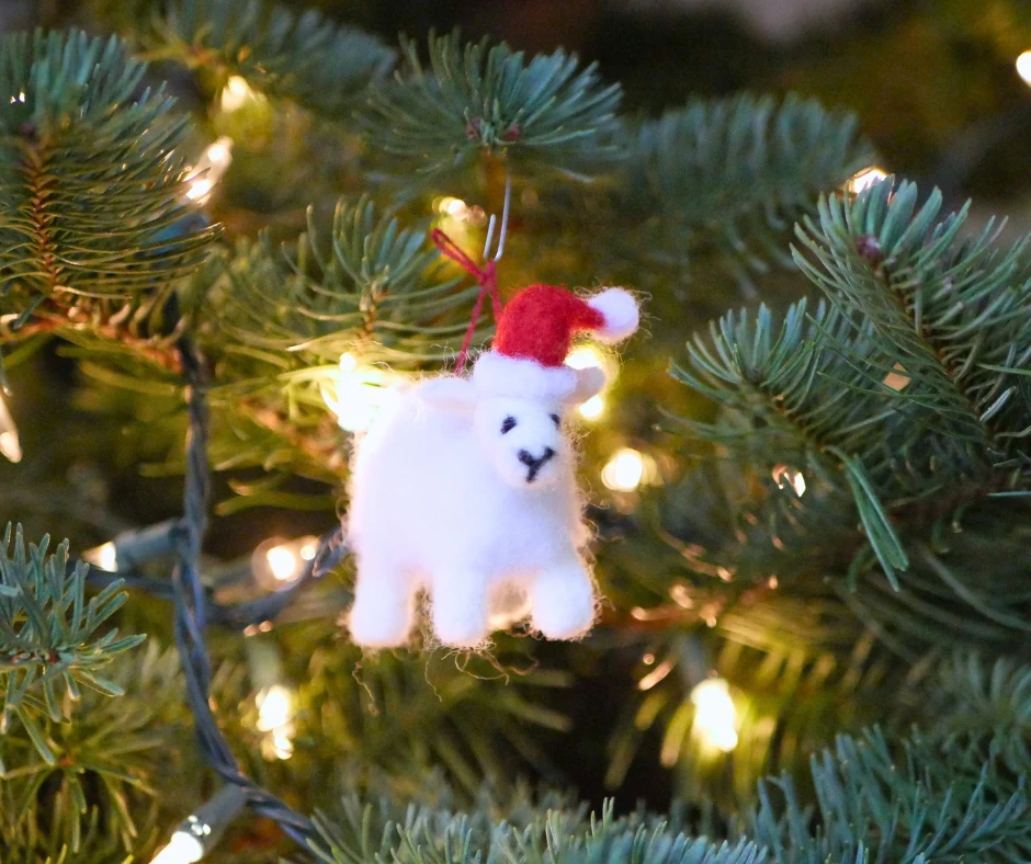 Image of a needle felted sheep wearing a Santa style hat hanging on an evergreen tree with white string lights. 