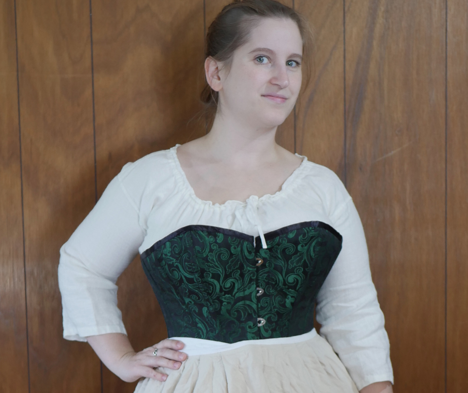 Image of a woman standing in front of wood paneling wearing a white shift and a cream color skirt with a black and green brocade corset. 