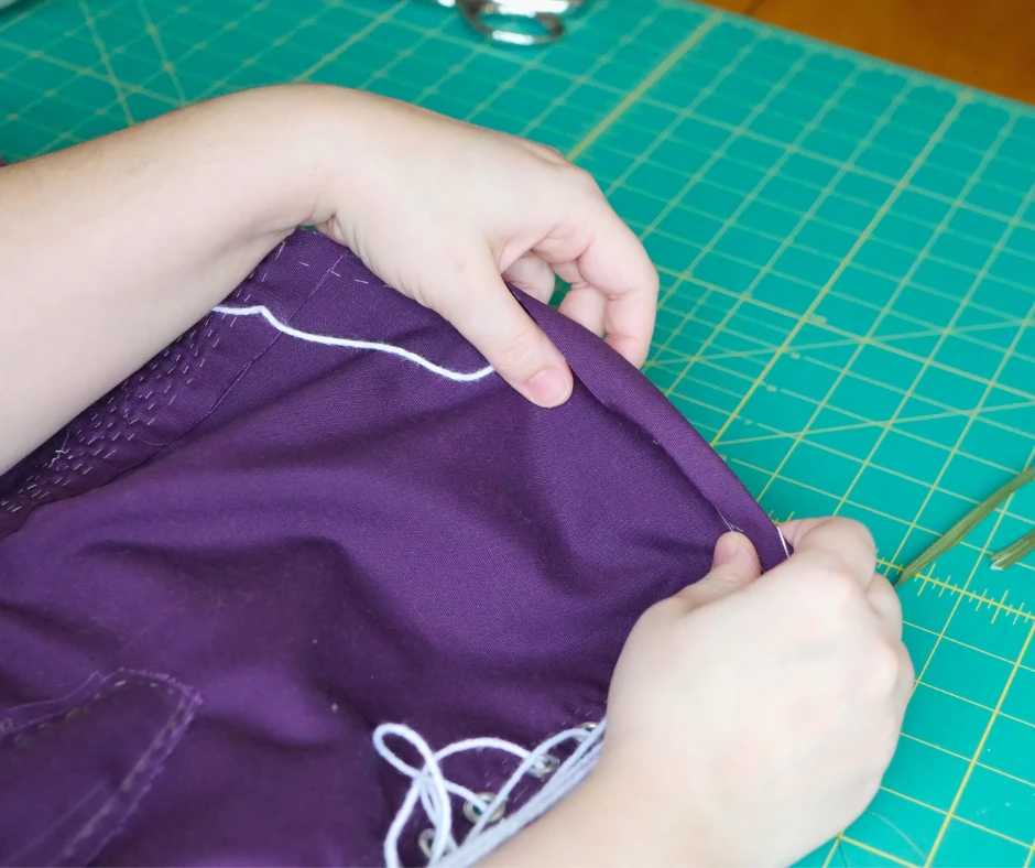 Image of a pair of white hands turning the edge of a binding over the top of a purple corset, being sure to tuck in the drawstring. 