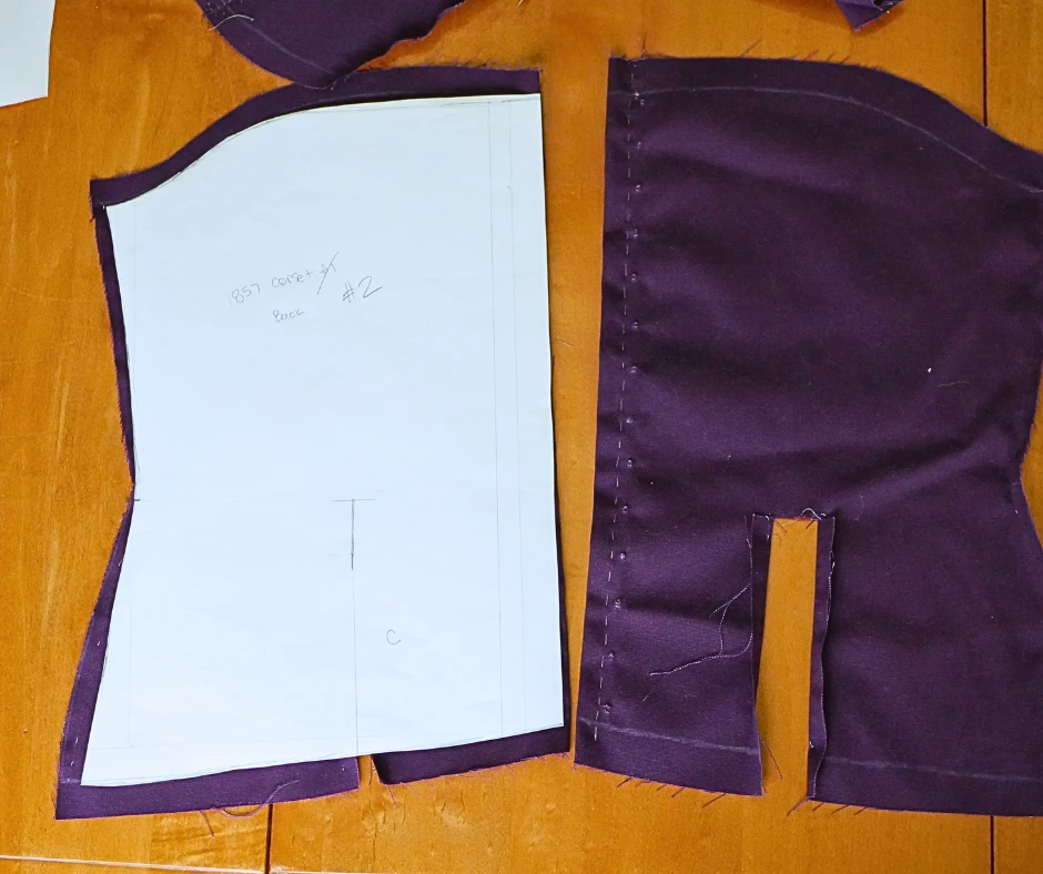Image of the back piece of a corset in purple fabric with a back pattern piece laying over the fabric to show the difference between the first pattern and the second pattern. 