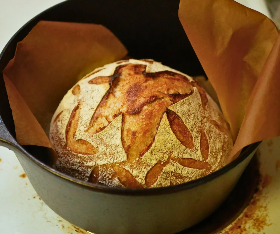 Image of a round loaf of freshly baked sourdough bread with decorative scoring in a cast iron dutch oven. 