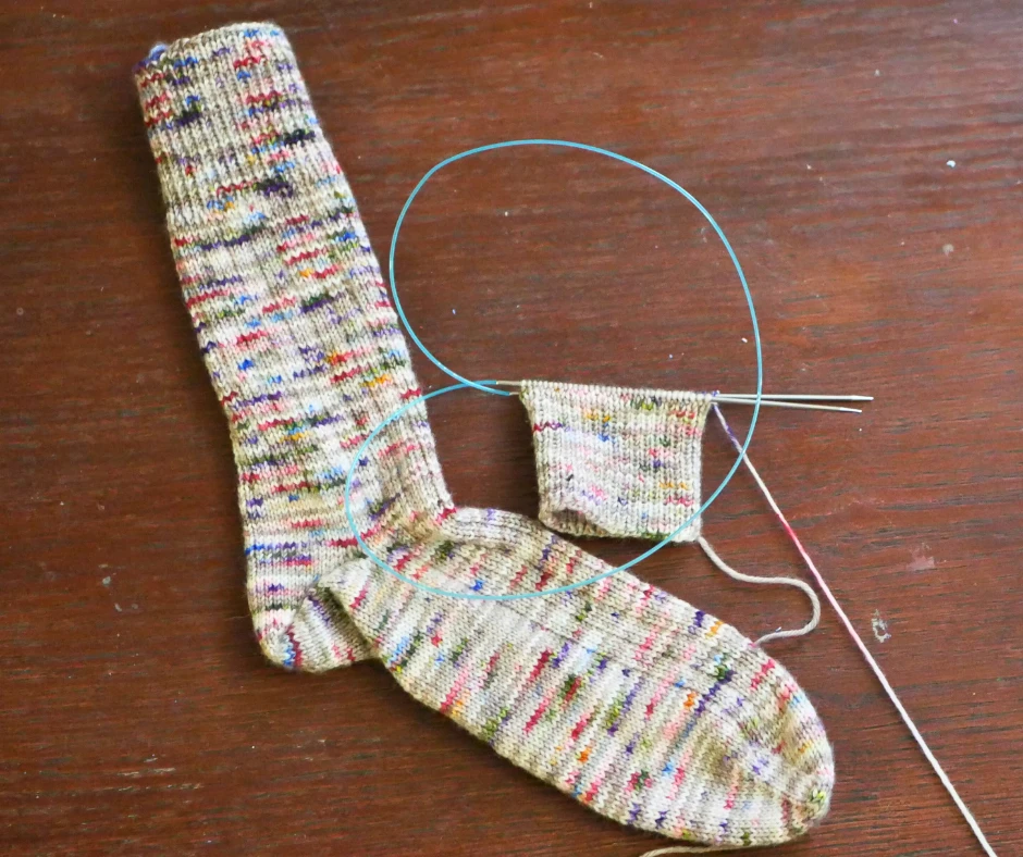 Image of a hand knit sock in brown yarn with pops of purple, blue, pink, orange, and green, with a partially knit sock in the same color, still on a pair of circular knitting needles with a blue cable. 