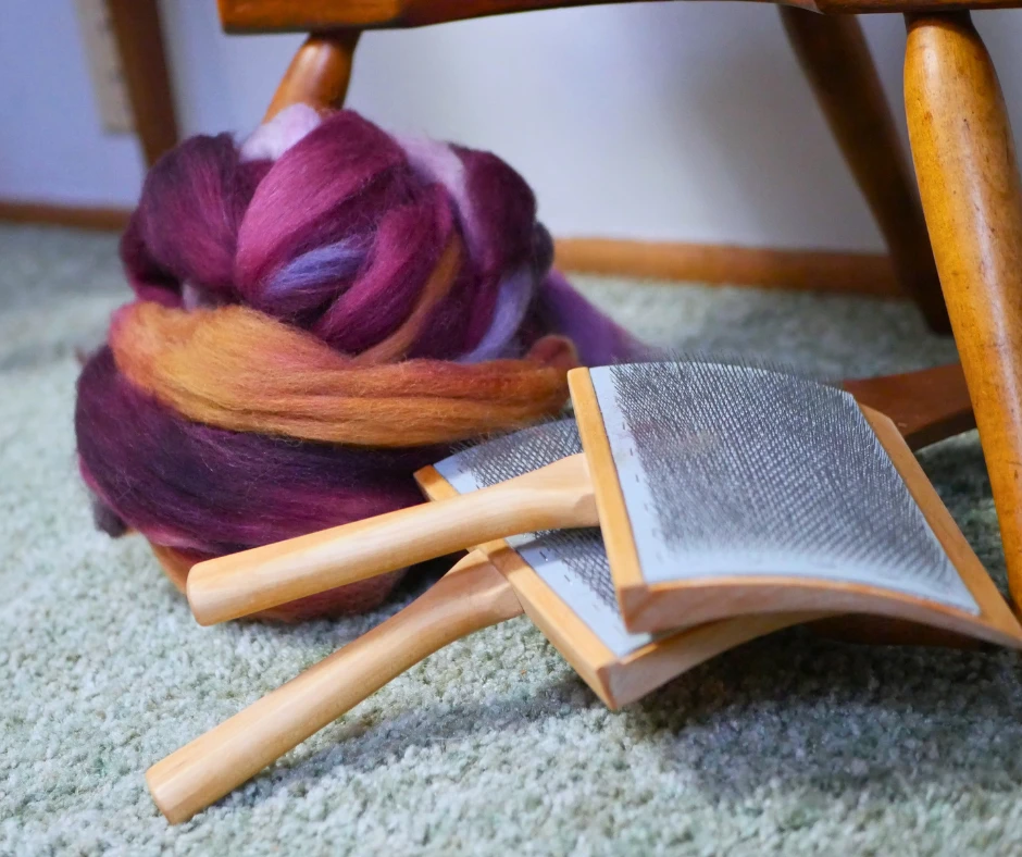 Image of a nest of purple and orange fiber for spinning, resting near the feet of a spinning wheel, next to a pair of hand carders. 