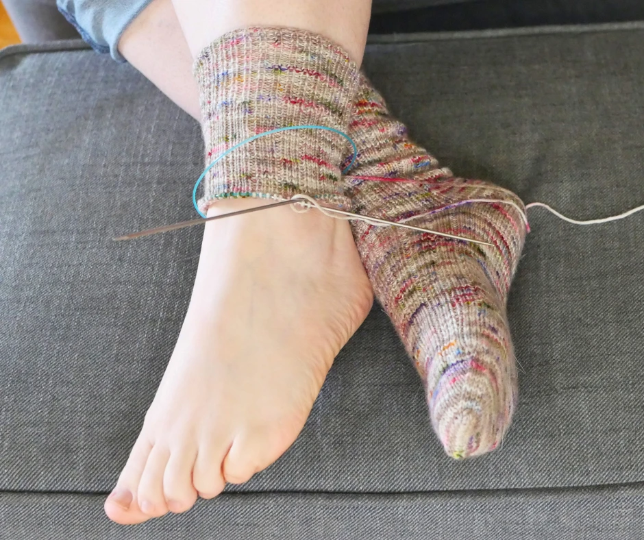 Image of a pair of crossed feet. The bottom foot has a knit sock in brown with pops of color. The top foot only has a knit ankle cuff, since the second sock is not yet finished being knit. 