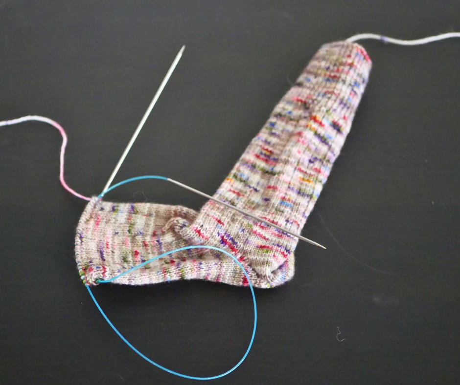 Image of a partially knit sock on a set of circular knitting needles with a blue cable. The sock yarn is brown with pops of blue, purple, green, orange, and pink. 