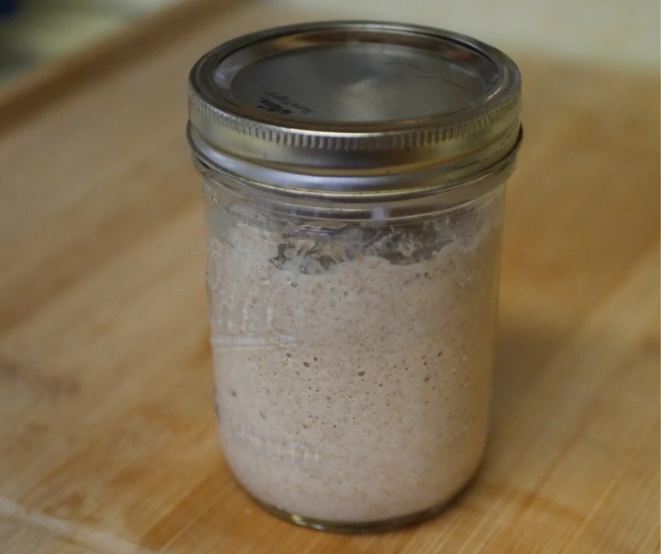 Image of a wide mouth mason jar sitting on a wood cutting board with the lid on. In the jar is a whole wheat sourdough starter full of bubbles. 