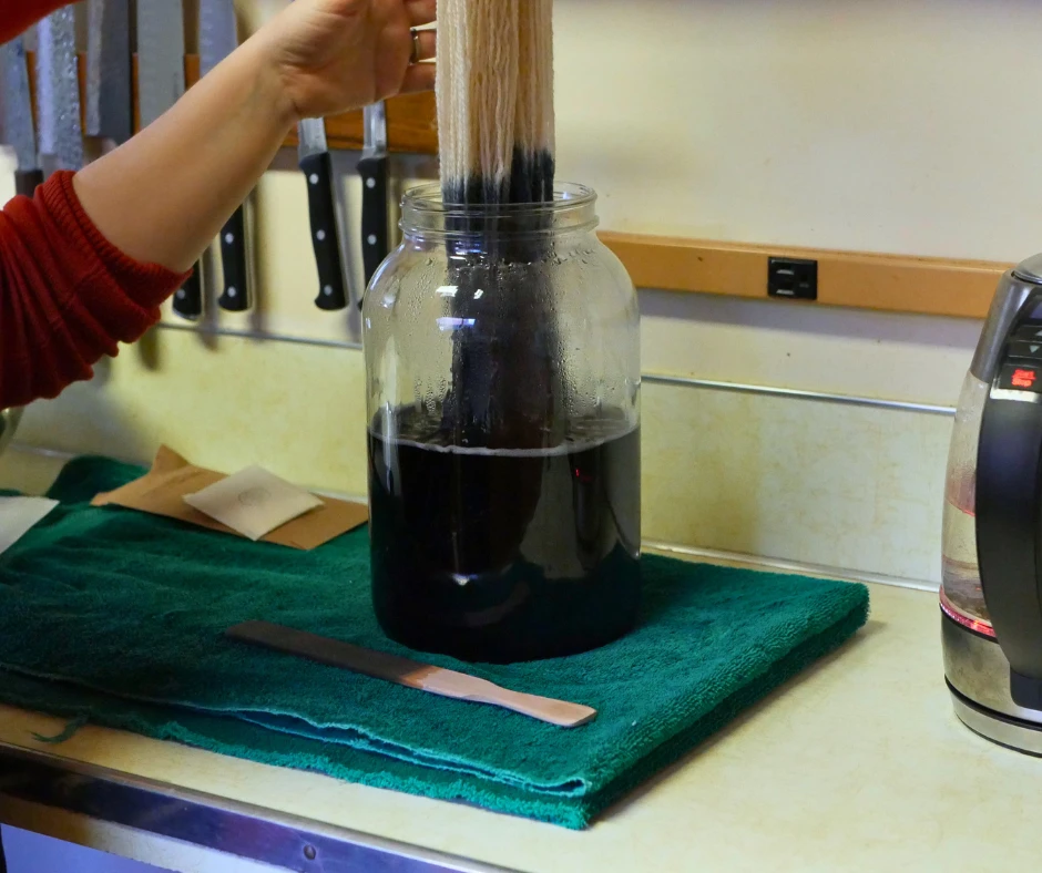 Image of a large glass jar half full of dark liquid with yarn being placed in it to dye. 