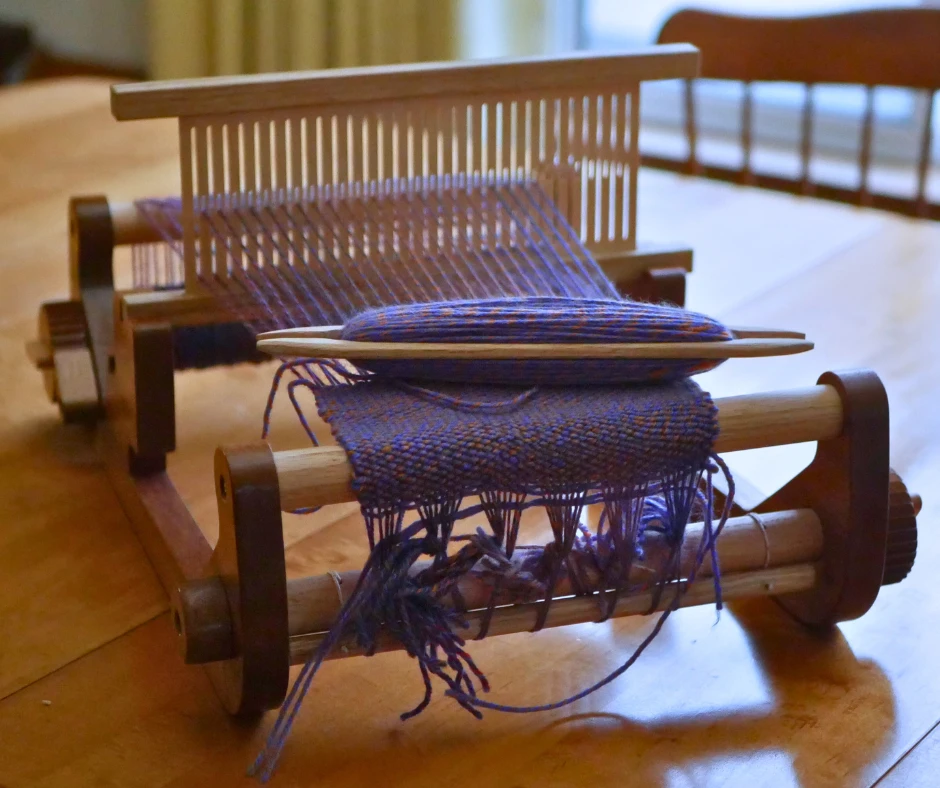 Image of a wooden table rigid heddle loom with blue warp and weft. A shuttle full of yarn rests across the fabric, ready to weave.  