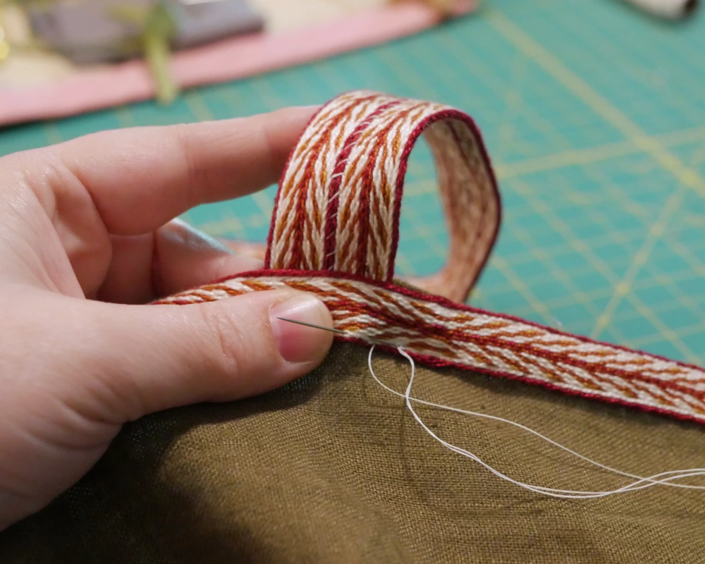 Finishing the raw edges of the top of a Viking apron dress by covering it with some hand woven trim. 