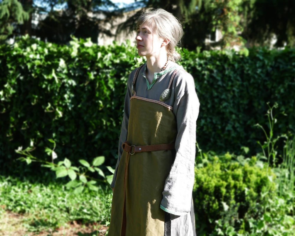 A white woman stands in front of a wall of ivy, wearing a green Viking apron dress over a gray linen tunic. 