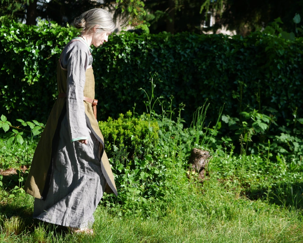 A woman walks through a field of green wearing a Viking inspired outfit. The bottom layer is a gray linen dress with long sleeves and skirt, with a green trim at the neckline and the end of the sleeves. Over that is a green apron dress with oval broaches and a brown leather belt. 