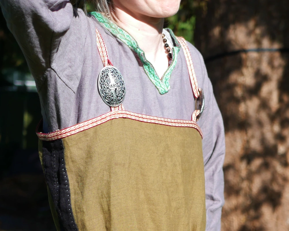 Close up of a green linen Viking apron dress fastened with large oval broaches made of pewter. 