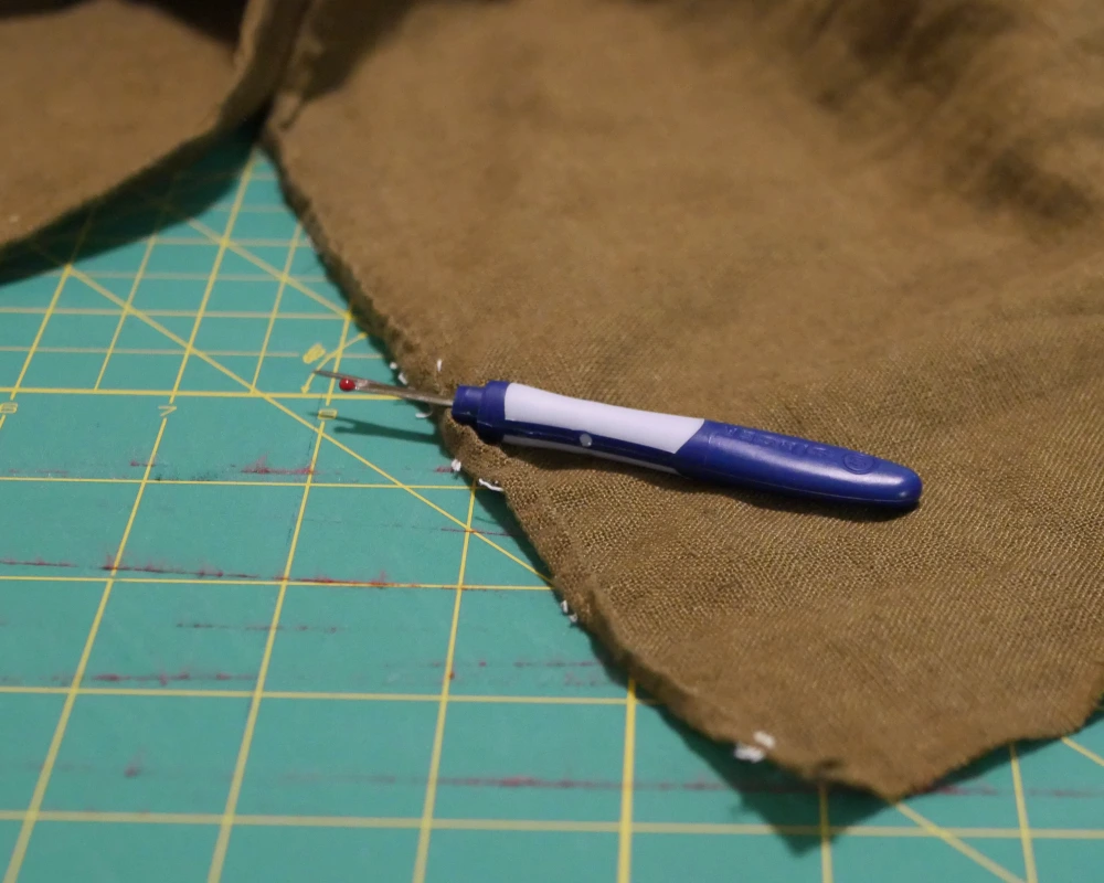 Close up of a blue seam ripper resting on green linen fabric that has recently had a seam removed. 