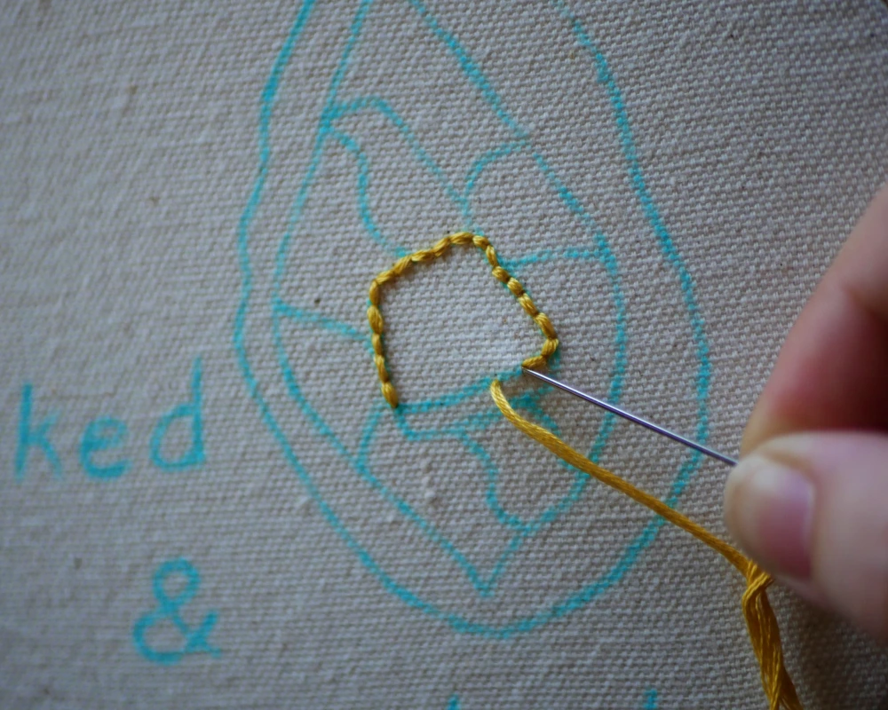 A close up of a back stitch outline of a pat of butter being stitched with yellow embroidery floss.