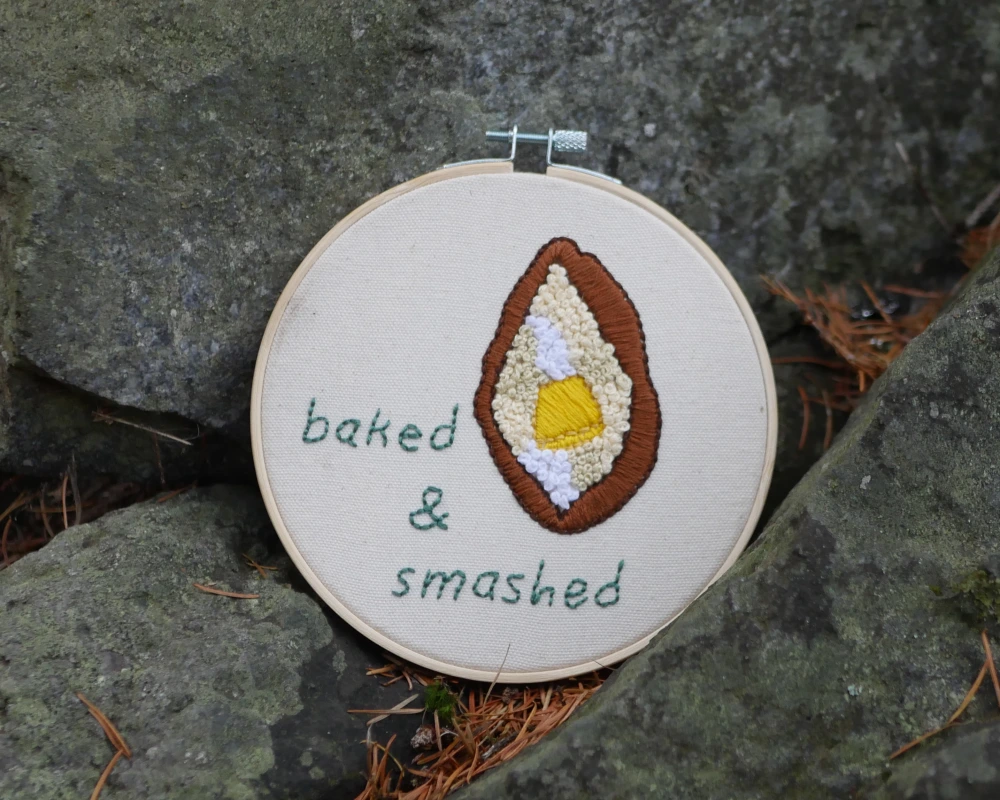 An embroidery hoop rests on large gray rocks. Fabric embroidered with the words, "baked and smashed" and an image of a baked potato from the top down, so that it is an open, elongated oval. A yellow pat of butter in the middle completes the image.