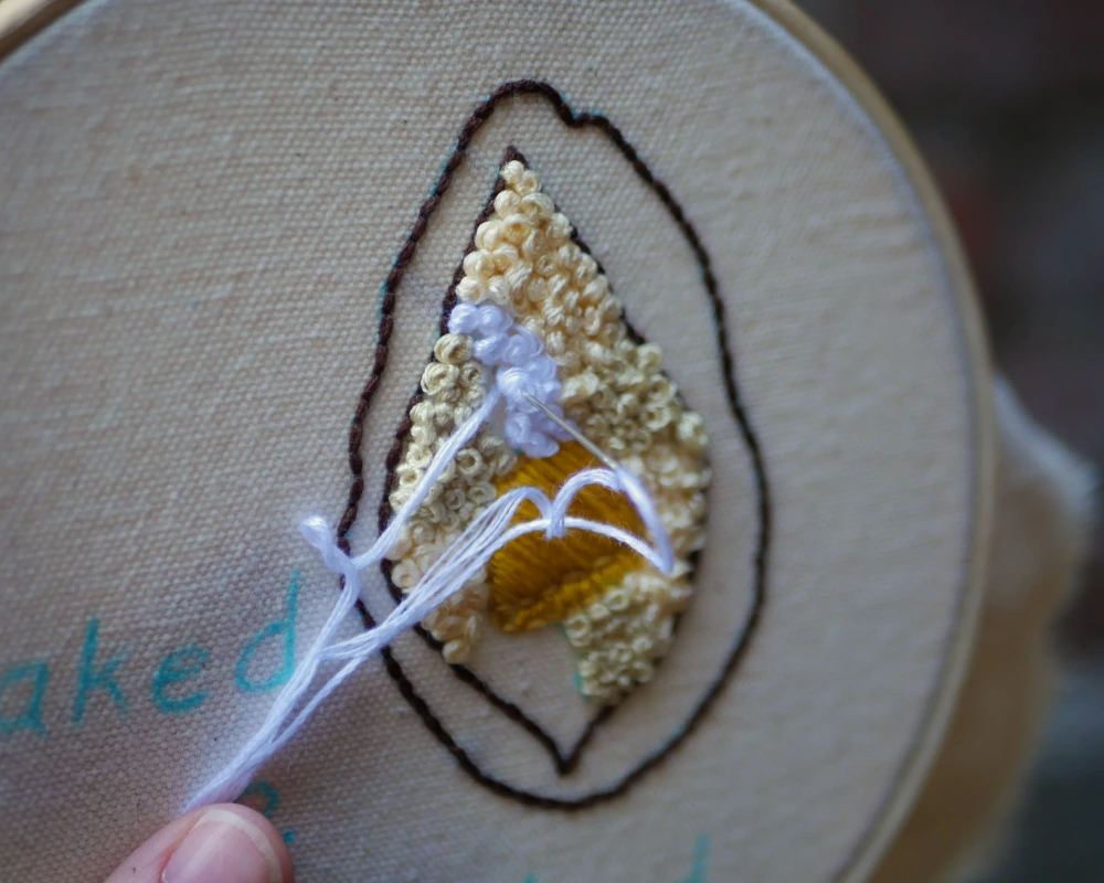 Sewing french knots to create the texture of the inside of a baked potato on an embroidery piece.