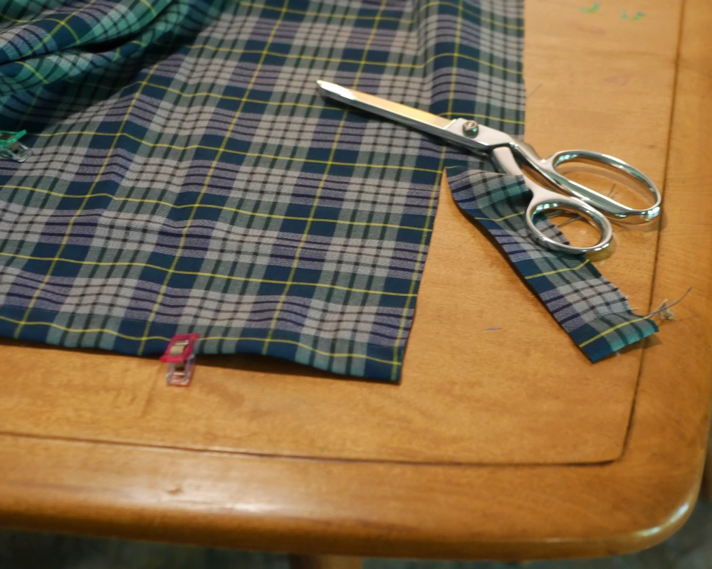 The bottom edge of a plaid curtain with a large pair of fabric shears showing the process of evening out the bottom hem of the curtain.
