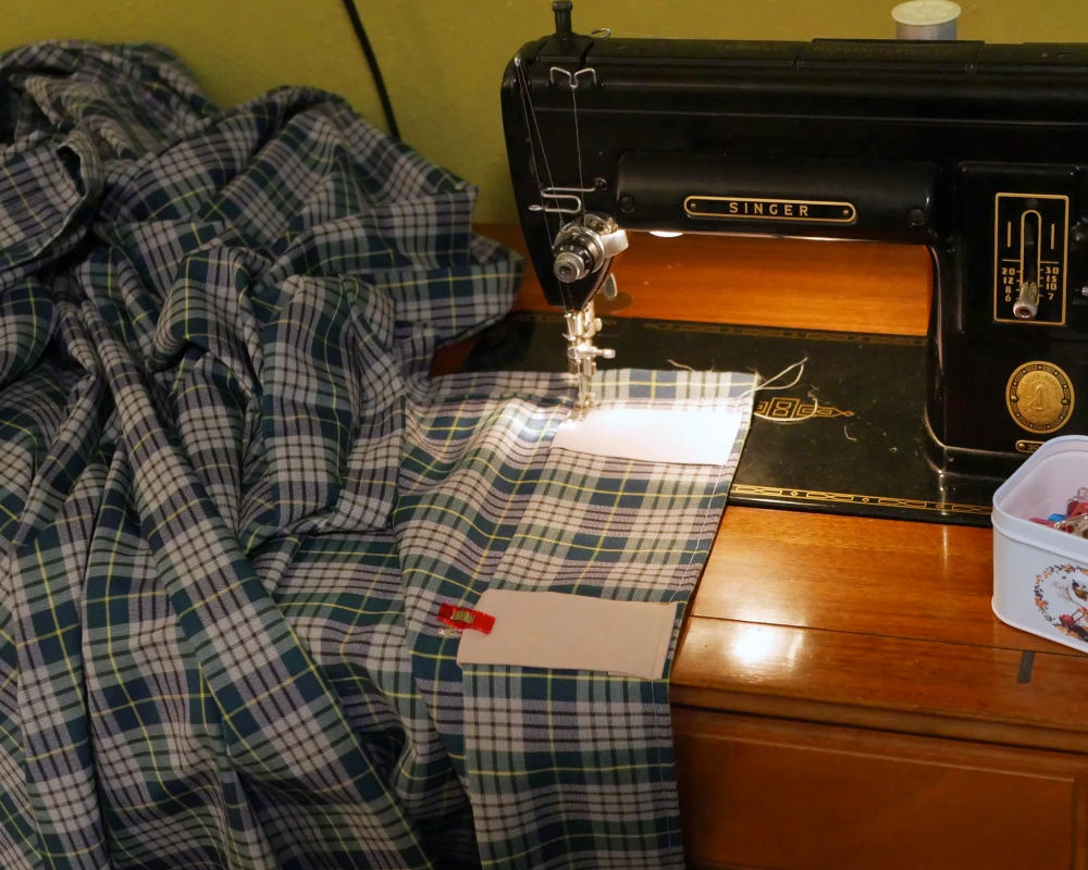 The top edge of a green and gray plaid curtain panel having the curtain tabs being sewn on by sewing machine. 