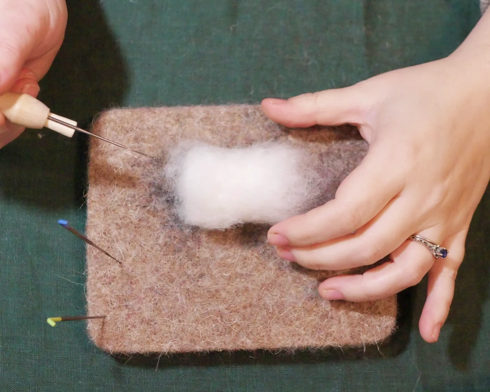 A pair of hands preparing to needle felt a ball of white wool.