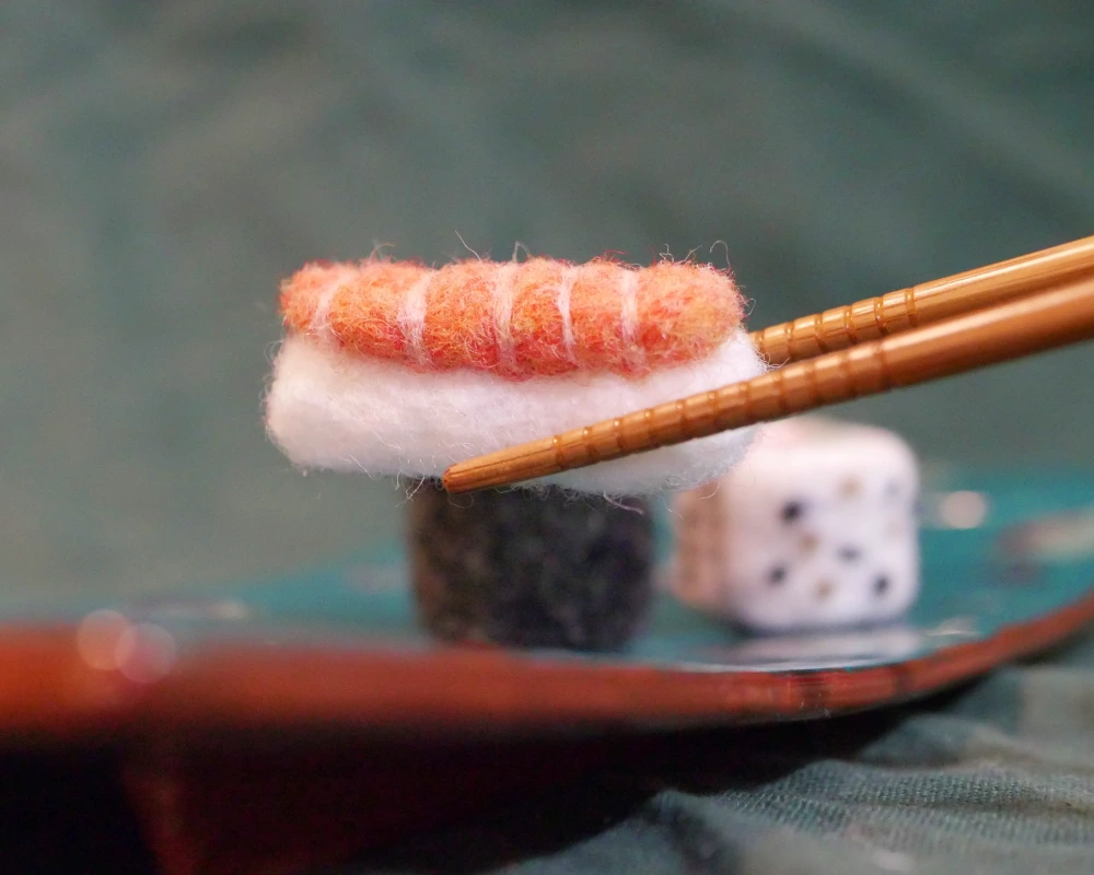 Close up of a small needle felted sculpture of a salmon nigiri is held by a pair of wooden chopsticks.