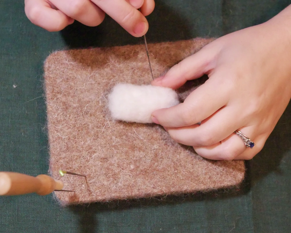 White wool that has been felted into a tubular shape being held by one hand as a felting needle is positioned above it.
