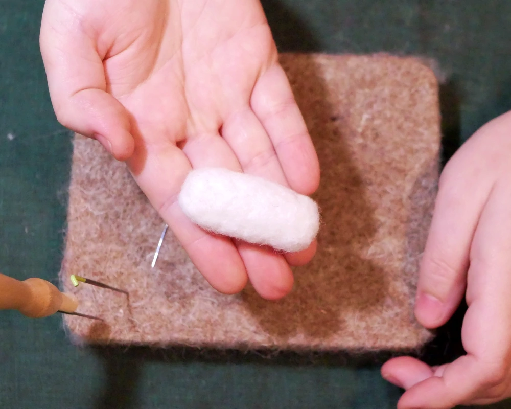 A hand with palm up holds a small tubular shaped piece of white wool that has been needle felted.