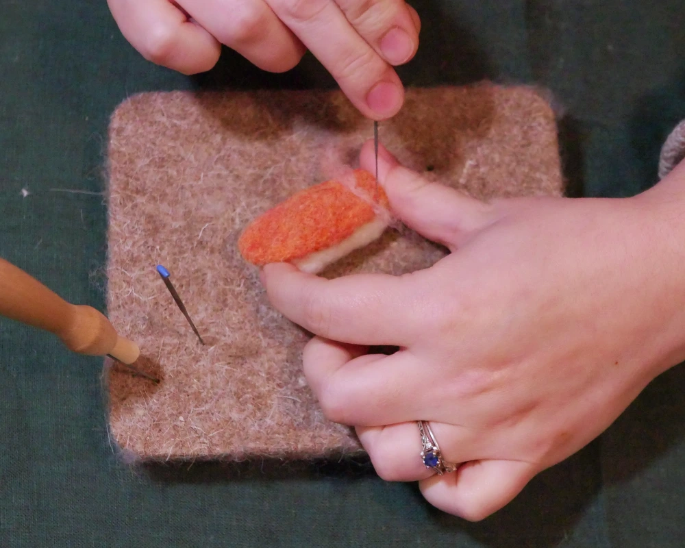 Thin strips of pink wool are being worked into a needle felted sculpture of salmon nigiri.