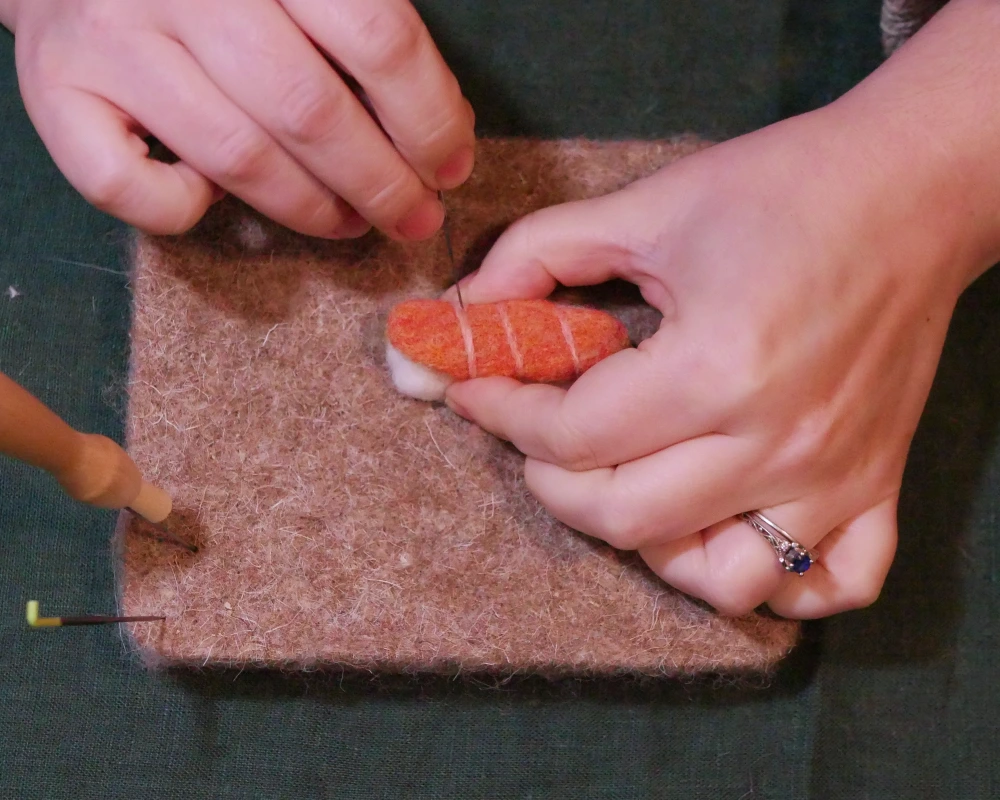 Thin strips of pink wool are being worked into a needle felted sculpture of salmon nigiri.