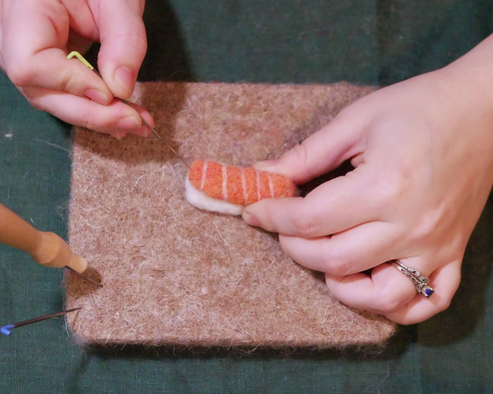A fine felting needle being used to for finishing details on a salmon nigiri needle felted sculpture.